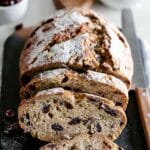 A rustic loaf of cranberry walnut bread, dusted with flour and sliced on a dark slate board.