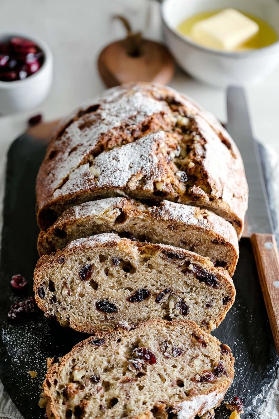 cranberry-walnut-bread A rustic loaf of cranberry walnut bread, dusted with flour and sliced on a dark slate board.