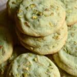 A close-up of a plate piled high with soft, pale green pistachio pudding cookies, with visible chunks of chopped pistachios on top.