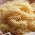 A close-up of fresh, uncooked homemade kataifi dough, showing its distinctive thin strands resting in a parchment-lined basket.