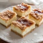Four golden-brown Mexican cheesecake bars arranged on a white decorative plate, with a visible flaky crust, creamy filling, and cinnamon-sugar topping