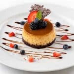 A beautifully presented mini chocoflan on a white plate, featuring a glossy caramel top, creamy flan center, and dark chocolate cake base, garnished with fresh blackberries, blueberries, a strawberry slice, mint, a decorative chocolate piece, and chocolate drizzle.