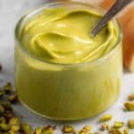 A close-up shot of a small glass jar filled with creamy, vibrant green pistachio cream. A small spoon is dipped into the cream, and shelled pistachios are scattered around the base of the jar on a light-colored surface.