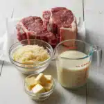 Ingredients for steak fettuccine alfredo laid out on a white wooden surface: two raw New York strip steaks on parchment paper, a glass bowl of grated Parmesan cheese, a measuring jug of heavy cream, and a small glass bowl of butter cubes